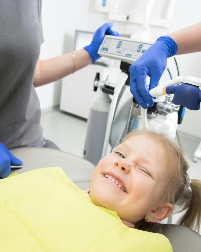 A cheerful child winks while sitting in a dental chair, surrounded by dental professionals.